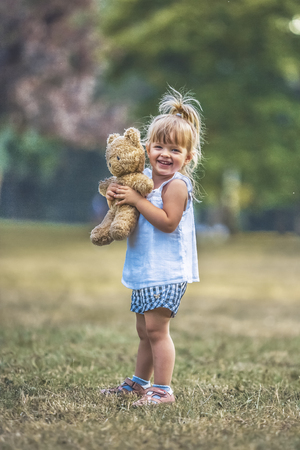 Little beautiful blonde girl holding teddy bear toy in her arms on a grass field during summer day.の写真素材