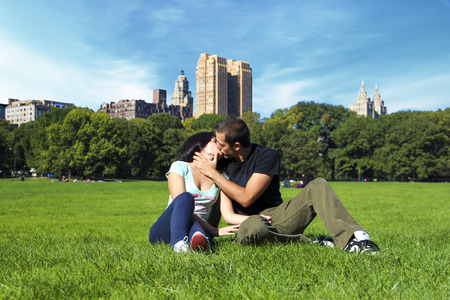 Cute couple kissing in Central Park New York City on a grass field with Manhattan buildings in background during sunny summer dayのeditorial素材