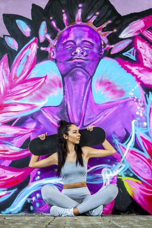 Young fit beautiful girl sitting outdoor with skateboard on her shoulders and street graffiti in the background during summer sunny dayの写真素材