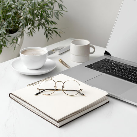 Laptop, coffee cup, notebook and glasses on white table.