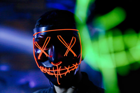 A man is seen wearing a brightly colored neon mask inside a poorly lit room.の写真素材