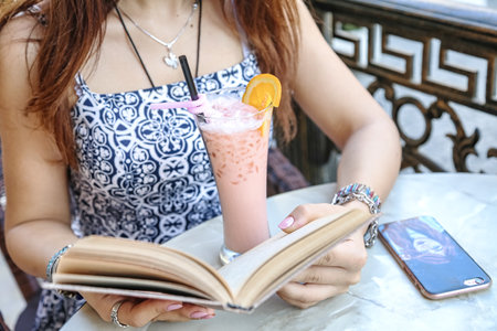 A woman is seated at a table, engrossed in a book while sipping on a drink.の写真素材