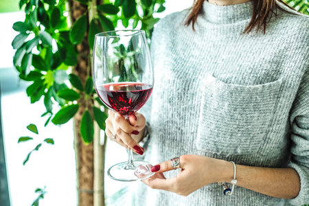 A woman dressed in a stylish outfit holds a wine glass filled with red wine, enjoying a...