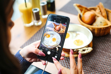 A woman leans over a plate of food, focused on capturing the perfect picture with her mobile phone.の写真素材