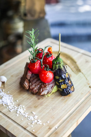 A piece of succulent steak accompanied by ripe tomatoes and an assortment of fresh vegetables neatly arranged on a wooden cutting board.の写真素材