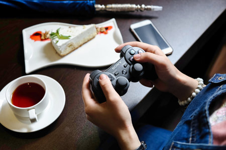 A person holds a video game controller while sitting next to a cup of tea.