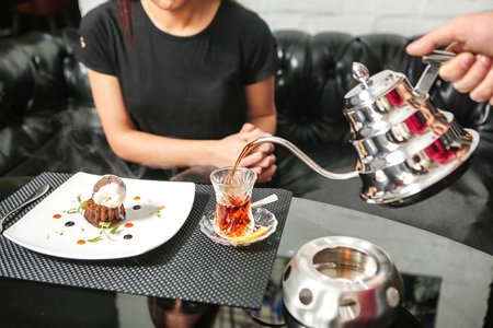 A woman carefully pours tea from a teapot into a glass on top of a table.