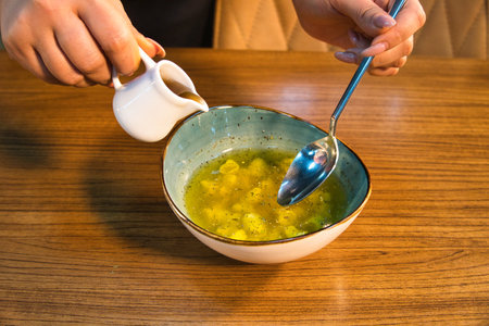 A person is pouring liquid from a small white pitcher into a bowl of soup. A spoon sits in the bowl.の写真素材