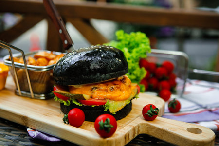 A black burger with cheese, lettuce, and tomato is served on a wooden board with fries and cherry tomatoes.の写真素材