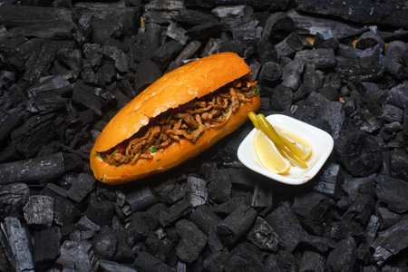 A grilled meat sandwich sits on a bed of charcoal with a side of lemon and peppers.の写真素材