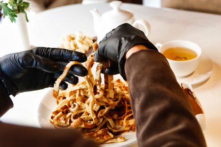 A person wearing black gloves is eating fried noodles with a fork from a white plate.