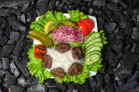A white plate with grilled meat, rice, lettuce, cucumbers, tomatoes, onions, and pickles sits on a bed of black charcoal.の写真素材