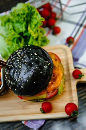 A close-up of a black bun burger with cheese and tomato on a wooden serving board.の写真素材