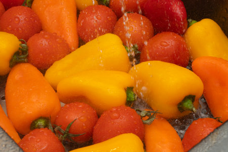 Colorful bell peppers and cherry tomatoes are washed in a kitchen sink, water cascading over them. This preparation hints at a healthy meal ahead.の写真素材