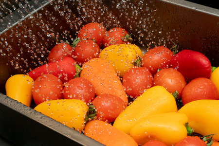Colorful cherry tomatoes and mini bell peppers are being rinsed under running water in a sink,...