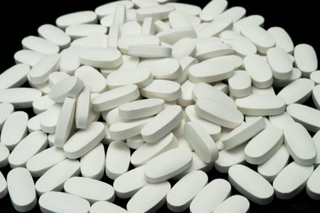 Numerous white oval tablets are spread across a shiny black surface, showing their uniform shape and...