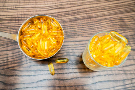 Fish oil capsules are arranged in a measuring cup and a glass on a wooden table. The bright yellow...