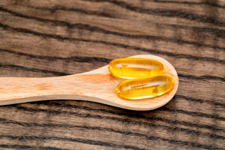 Two golden gel capsules sit on a small wooden spoon, placed on a rustic, dark wooden surface, emphasizing health and wellness themes.の写真素材