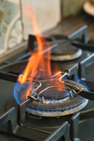 A vibrant blue and orange flame dances on a gas stove burner while cooking in a cozy kitchen filled with homey decor and warm lighting.の写真素材