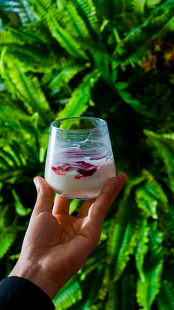 A hand presents a refreshing drink with rose petals, set against vibrant green foliage, showcasing a moment of relaxation.の写真素材