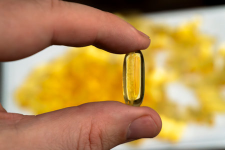 A hand grasps a large yellow gelatin capsule, with a blurred background of multiple similar capsules scattered on a surface.の写真素材