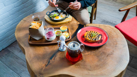 A person sits at a rustic wooden table savoring a delightful brunch spread featuring eggs, desserts,...