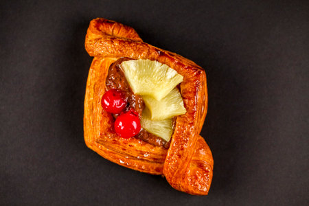 This pastry features a flaky crust topped with pineapple and cherries, richly glazed and presented on a dark background, inviting a delightful taste experience.の写真素材
