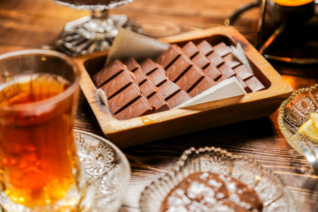 A warm drink sits beside a tray of neatly arranged chocolate bars on a wooden table, creating a cozy atmosphere for indulging in sweets and tea.の写真素材