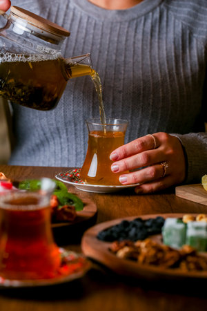 A person pours a warm drink into a glass while seated at a wooden table adorned with an array of colorful snacks and dishes, creating a cozy atmosphere.の写真素材