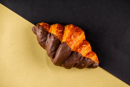A flaky croissant half-dipped in rich chocolate rests on a uniquely divided black and beige background, showing its glossy texture and golden layers.の写真素材