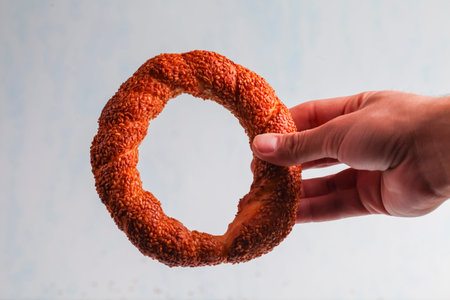 A hand holds a circular sesame-covered bread ring against a light, neutral background. The texture...