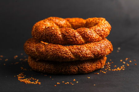 Three golden sesame rings are stacked neatly on a dark surface, while sesame seeds surround the...