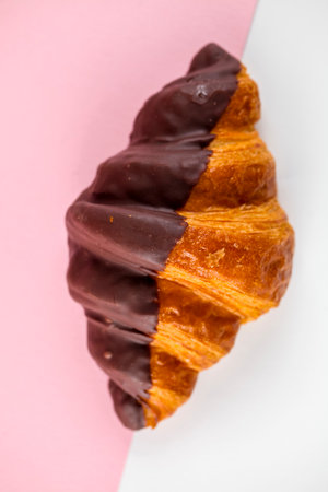 A freshly baked croissant is half-covered in rich chocolate, showing a golden, flaky texture. The background features soft pink and white hues, enhancing the visual appeal.の写真素材