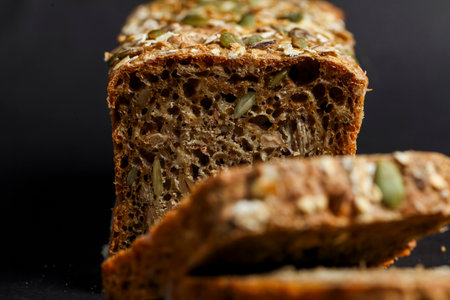 A loaf of whole grain bread made with various seeds is sliced, revealing its dense texture and rich color against a dark backdrop, highlighting its freshness.の写真素材