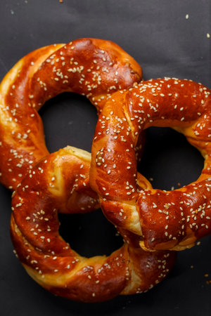 Three golden-brown pretzels are placed on a black surface, showing their shiny crust and sesame seed topping. Perfect for snacking or sharing.の写真素材