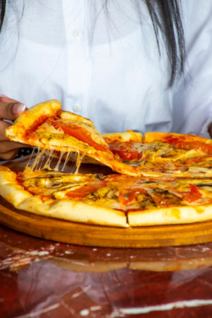 A person is pulling a slice of cheesy pizza from a round wooden platter, revealing melted cheese and toppings, in a warm dining atmosphere.の写真素材