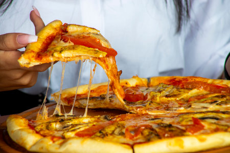 A person pulls a slice of pizza from a large platter, showcasing melted cheese and colorful toppings in a casual dining setting surrounded by friends.の写真素材