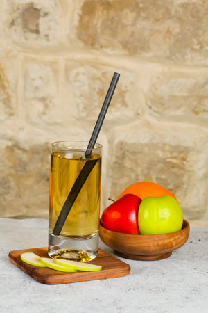 A tall glass filled with a golden beverage and a black straw stands alongside a bowl of colorful fruits including red, green apples and orange.の写真素材