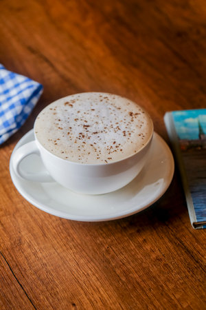 A creamy cup of coffee topped with froth sits on a saucer on a wooden table. A checkered cloth and an open magazine complement the setting.の写真素材