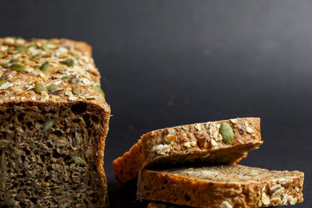 Slices of whole grain bread with seeds sit on a dark surface, showing a crusty exterior and a soft...