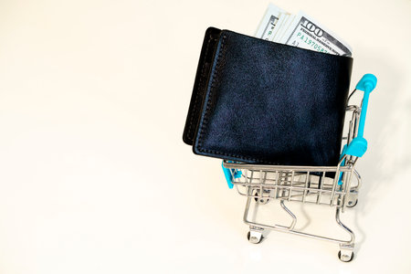 A black wallet sits in a miniature shopping cart filled with cash, highlighting the relationship between spending money and shopping culture at any time.の写真素材