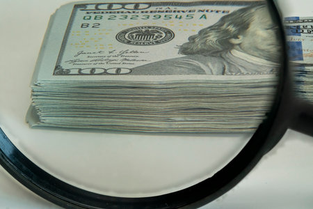A close-up view of neatly stacked hundred-dollar bills being closely inspected with a magnifying glass, highlighting details of the banknotes on a plain surface.の写真素材