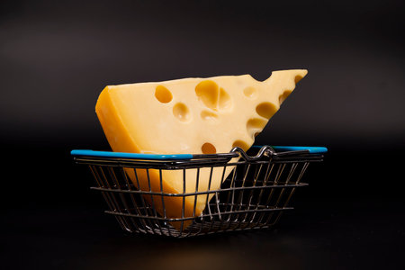 A wedge of cheese with characteristic holes rests in a small shopping basket. The dark background highlights the yellow color and texture of the cheese.の写真素材