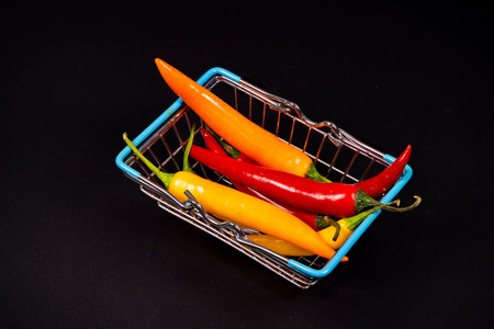 A small shopping basket holds an assortment of vibrant chili peppers, including red, yellow, and green varieties. The arrangement highlights their fresh colors.の写真素材