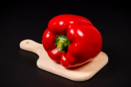 A bright red bell pepper sits on a wooden cutting board with a dark background, showing its shiny skin and vibrant color, ready for meal preparation.の写真素材