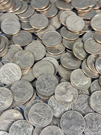 Many coins are spread out on a table, reflecting light. The scene shows the surface covered with...