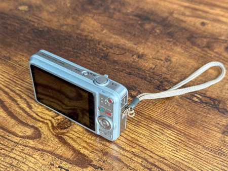 A digital camera sits on a wooden table. The camera shows a silver body and is positioned with its...