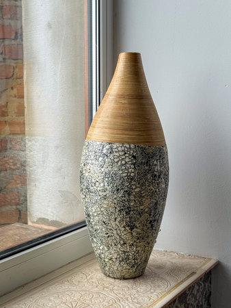 A tall vase sits on a window sill, with a narrow top made of wood and a wide bottom with a textured stone-like finish. Natural light shines on the vase from the window.の写真素材