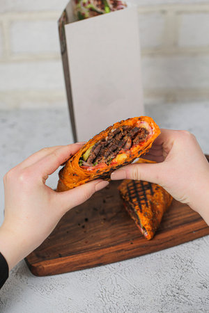 Two hands hold a stuffed flatbread filled with cooked meat and vegetables. A wooden board is in the background with the flatbread resting on it.の写真素材