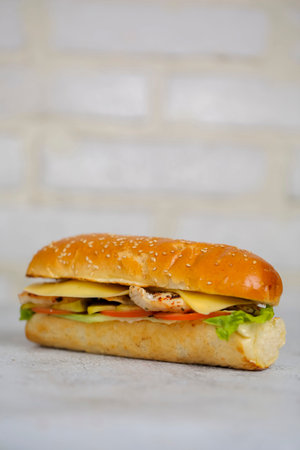 A sandwich sits on a flat surface. It has chicken, cheese, and vegetables inside a soft bun. The background is simple and light.の写真素材